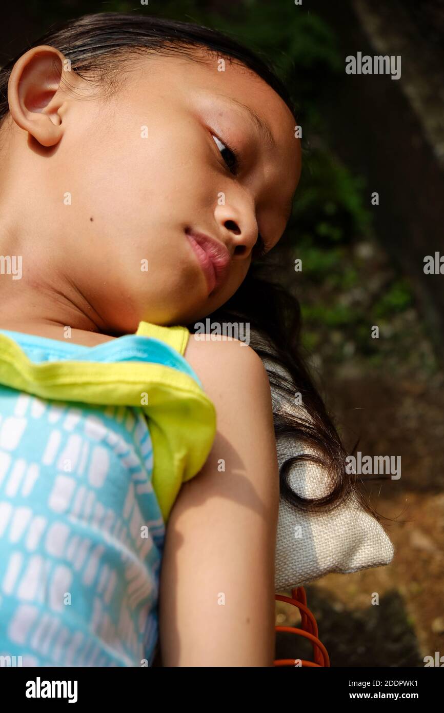 Nette asiatische Ethnizität kleines Mädchen auf einer Bank liegend, Sonnenbaden am Morgen zu Hause. Hand steigt auf, um ihr Gesicht von harten Sonnenlicht zu decken Stockfoto