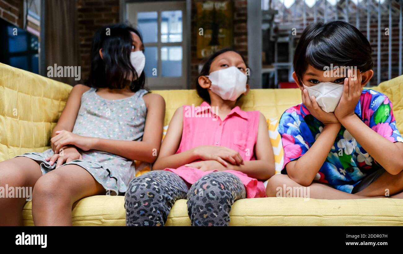 Südostasiatische Kinder tragen medizinische Gesichtsmaske auf Sofa sitzen Zusammen Fühlen Sich Während Der Isolation Zu Hause Gelangweilt Stockfoto