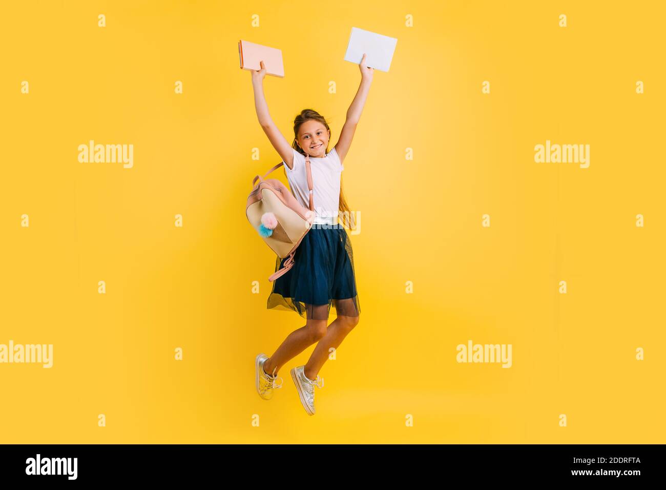 Glücklich Teenager-Mädchen in Schuluniform, Schülerin springt fröhlich mit Büchern auf einem gelben Hintergrund Stockfoto