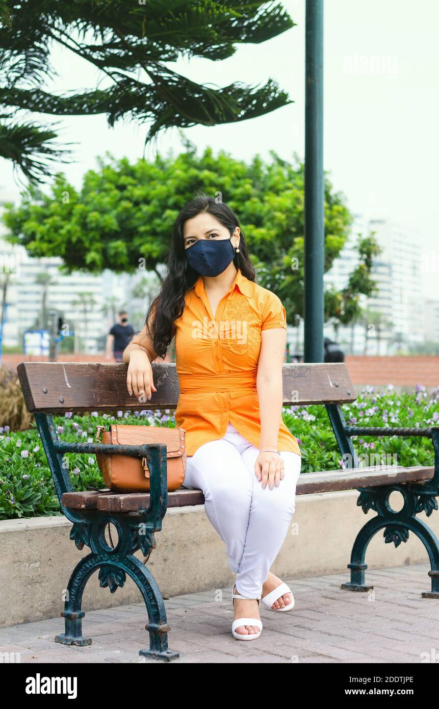 Frau trägt eine Maske, um sich vor dem covid19 sitzen in einem Park zu schützen. Frau, die sich im Frühling in einem Park entspannt und eine Schutzmaske trägt und während der Pandemie auf einer Bank in der Sonne sitzt Stockfoto