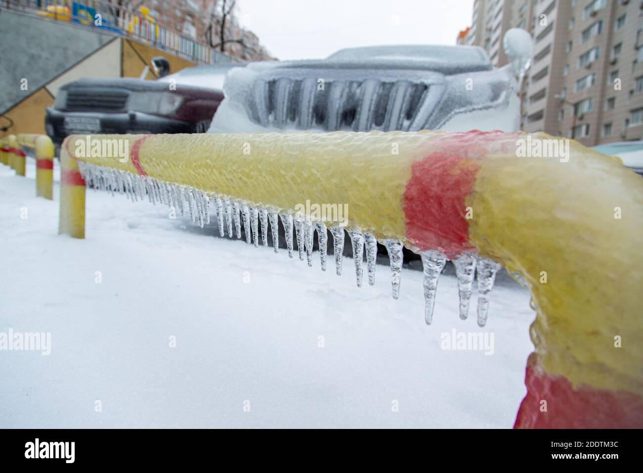 Gelb-roter Parkzaun in einer eisigen Kruste mit Eiszapfen. Vor dem Hintergrund der SUVs. Stockfoto