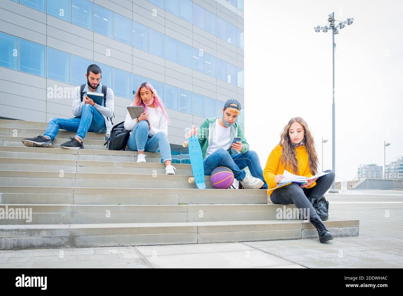 Junge Leute Universität studieng outoor während Coronavirus Pandemie. Vier multitnische Studenten mit Schutzmasken sitzen im Freien Stockfoto