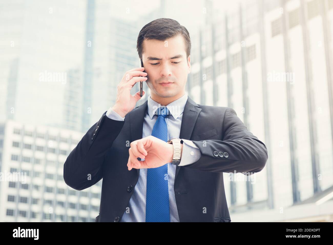 Geschäftsmann, der seine Uhr ansieht, während er anruft, um die Zeit zu überprüfen Handy im Hintergrund des Bürogebäudes verwacklungsunscharf Stockfoto