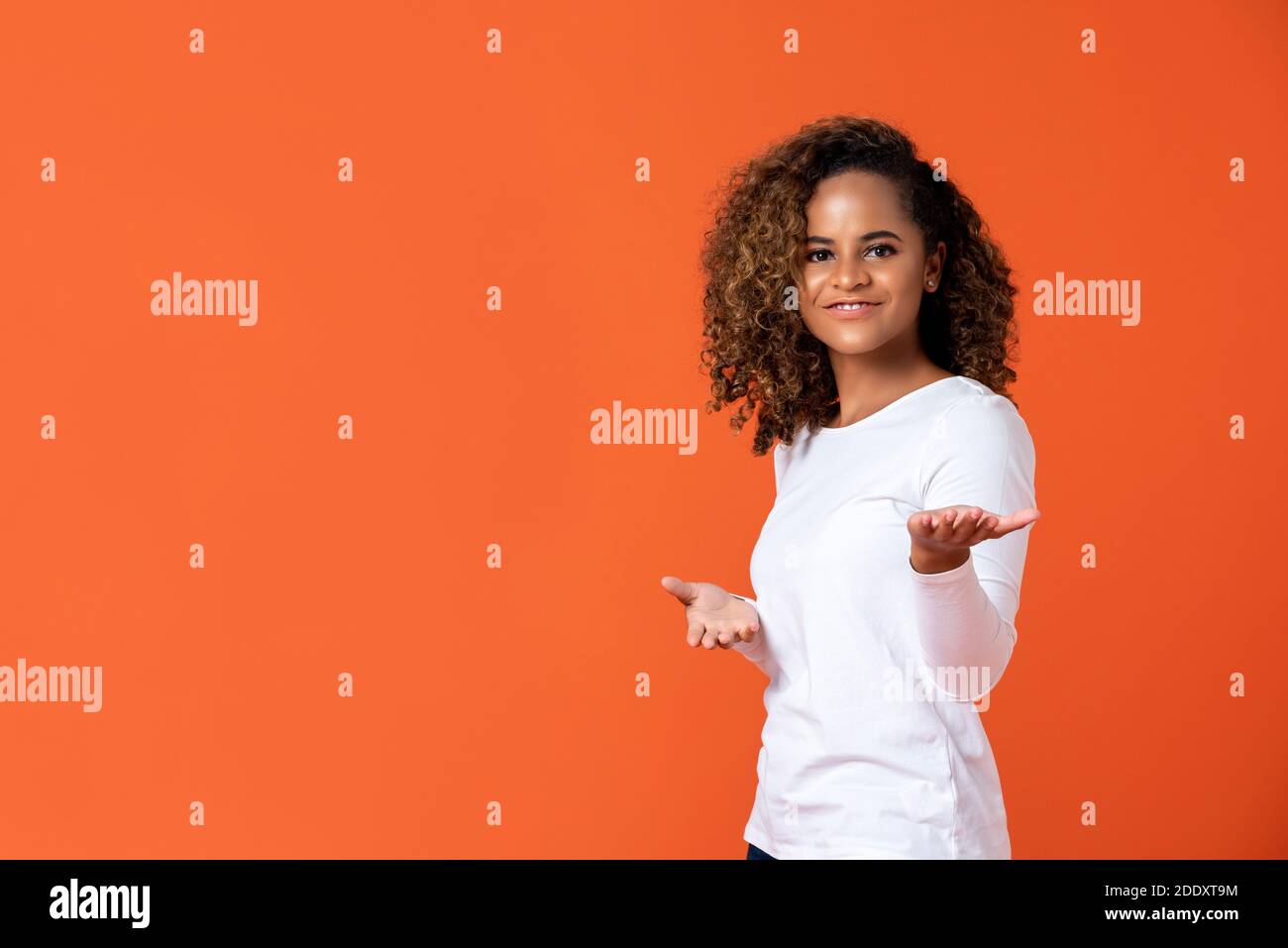 Glücklich lächelnde junge afroamerikanische Frau, die willkommen oder präsentiert Geste mit offenen Händen isoliert auf orangenen Studiohintergrund mit Speicherplatz kopieren Stockfoto