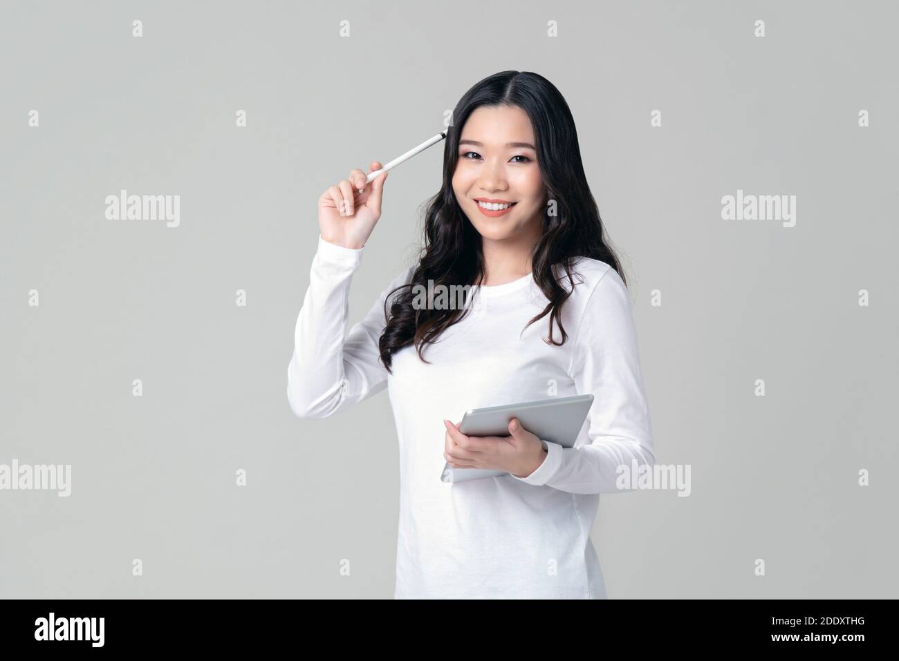 Lächelnde asiatische Frau in weißen Langarm-T-Shirt hält Stylus-Stift und Tablet in denken Geste isoliert auf hellgrauem Hintergrund. Stockfoto