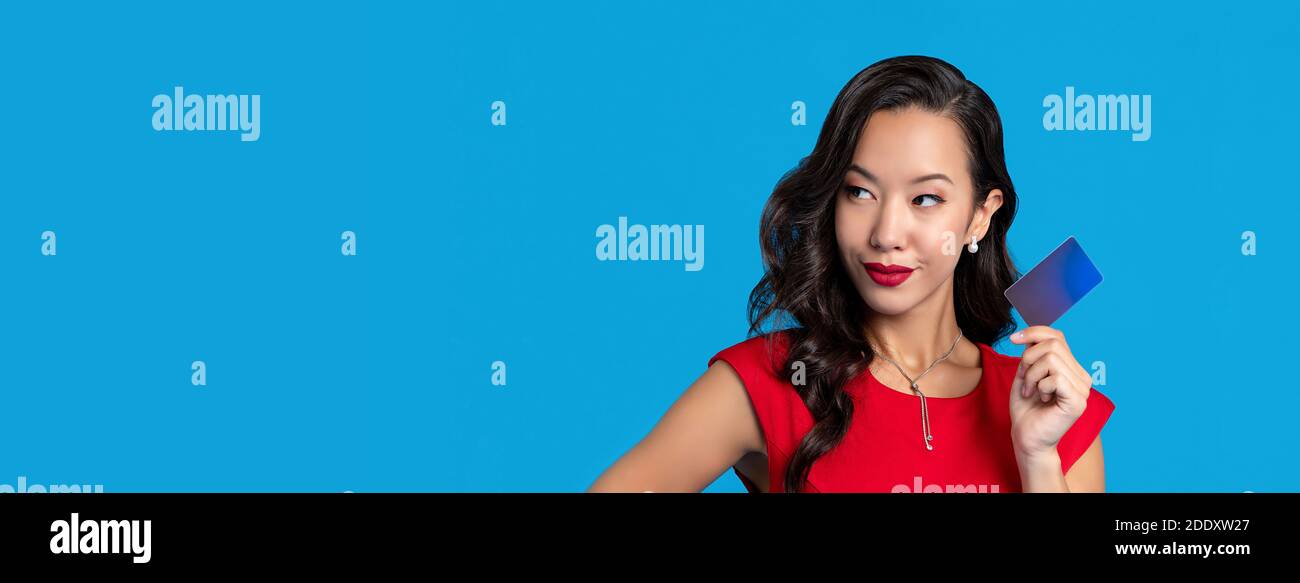 Wunderschöne gemischte asiatische Frau in rotem Kleid zeigt Kreditkarte In der Hand isoliert auf blauem Banner-Hintergrund mit Kopierraum Stockfoto