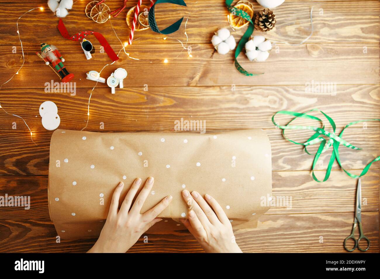 Draufsicht der Frauen Hände Verpackung Weihnachtsgeschenk auf Holztisch. Geschenkpapier mit weißen Punkten. Bänder Baumwolle Boxen trocken Orangen Girlande und Neujahr Spielzeug auf Hintergrund. Stockfoto