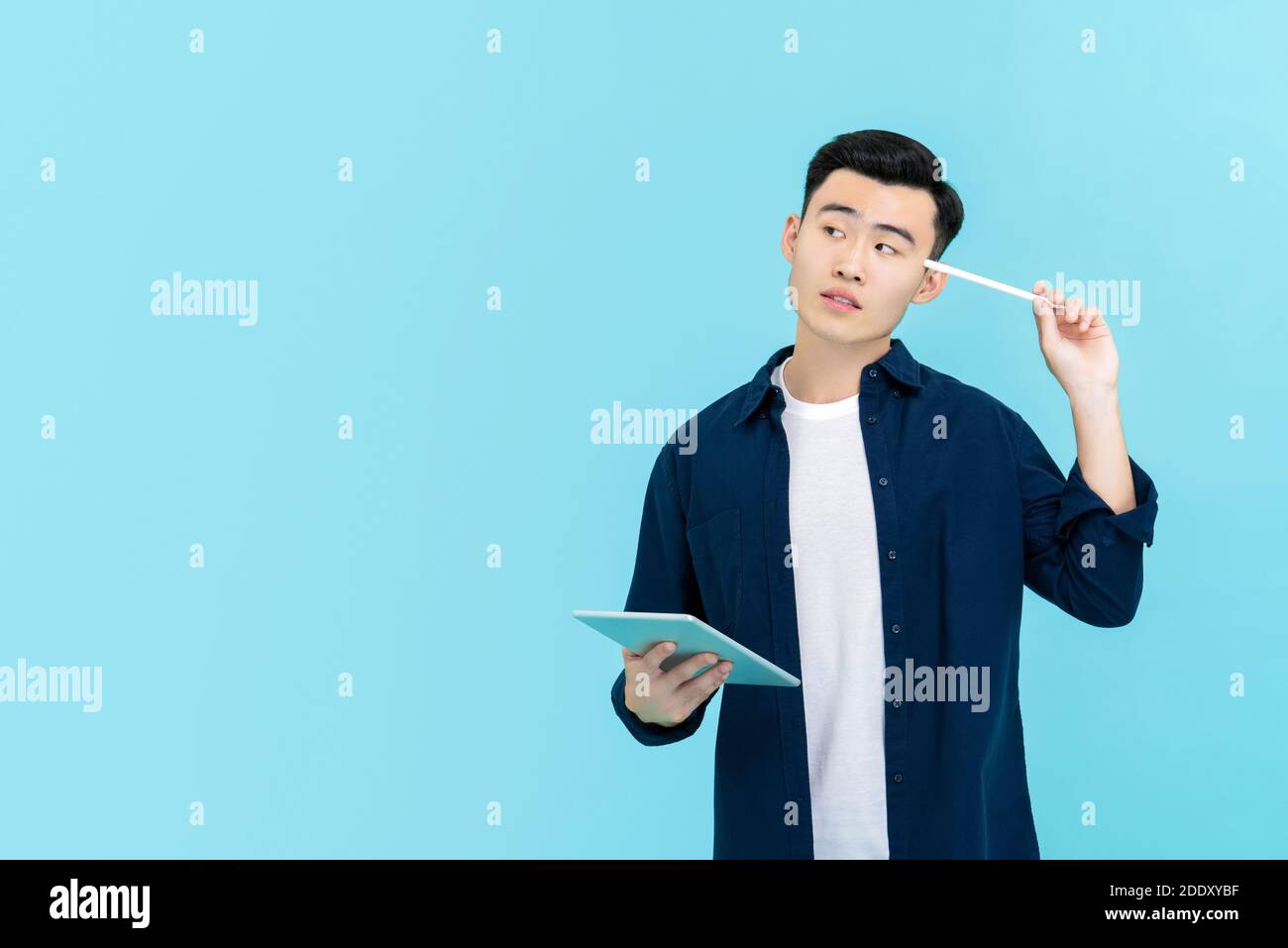 Junge gut aussehende asiatische Mann suchen nachdenklich und halten Stift zu Kopf mit Tablett in der Hand neben Kopierraum isoliert auf Hellblauer Hintergrund Stockfoto