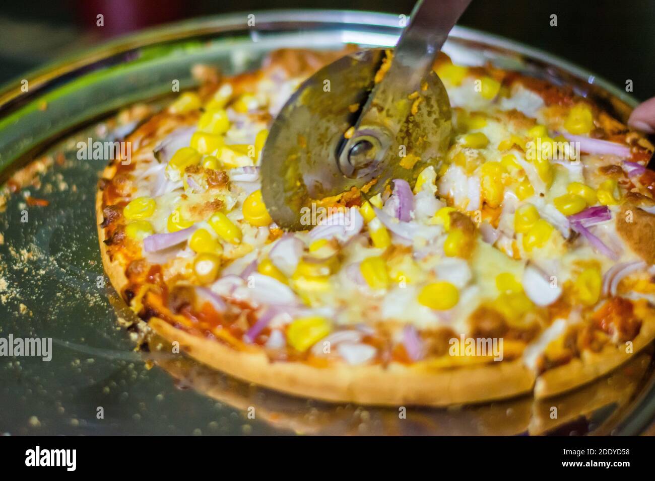 Schneiden von Käse Pizza mit Pizza Cutter, mit Tomaten, Zwiebeln, Mais Toppings, mit selektivem Fokus in Lockdown. Stockfoto