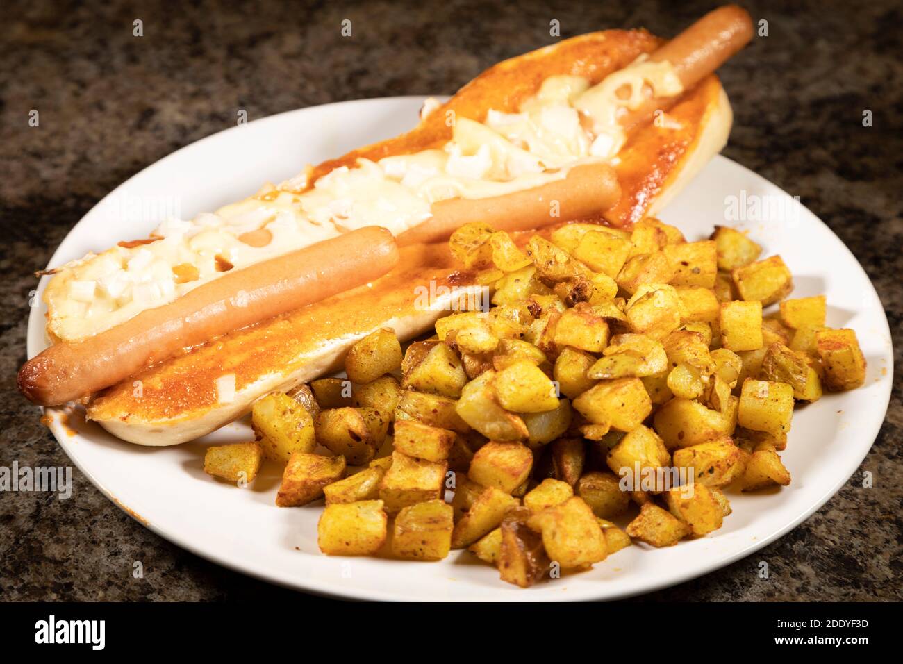 Ein spanischer Hot Dog serviert mit Patatas Bravas. Die Hot Dogs wurden in einem Baguette gebacken und mit Käse und gehackten Zwiebeln überbacken. Stockfoto