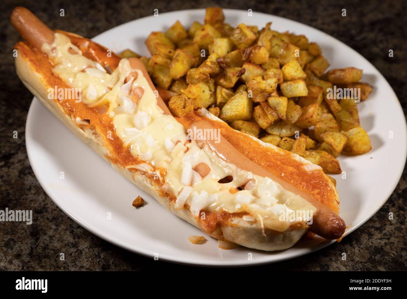 Ein spanischer Hot Dog serviert mit Patatas Bravas. Die Hot Dogs wurden in einem Baguette gebacken und mit Käse und gehackten Zwiebeln überbacken. Stockfoto