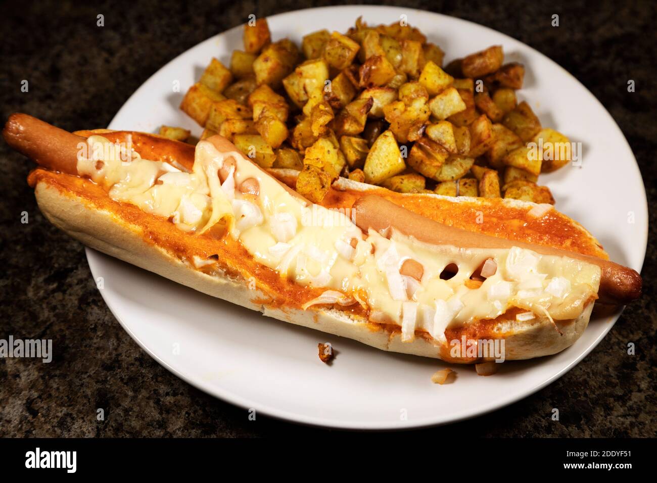 Ein spanischer Hot Dog serviert mit Patatas Bravas. Die Hot Dogs wurden in einem Baguette gebacken und mit Käse und gehackten Zwiebeln überbacken. Stockfoto