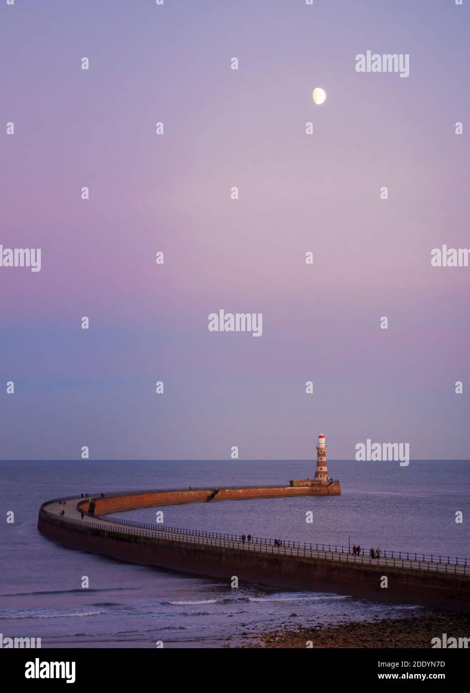Ein wunderschöner Sonnenuntergang am Roker Pier, Sunderland, Großbritannien, mit dem Leuchtturm und dem Mond im Blick und den Menschen, die sich am Pier entlang aufmachen. Stockfoto