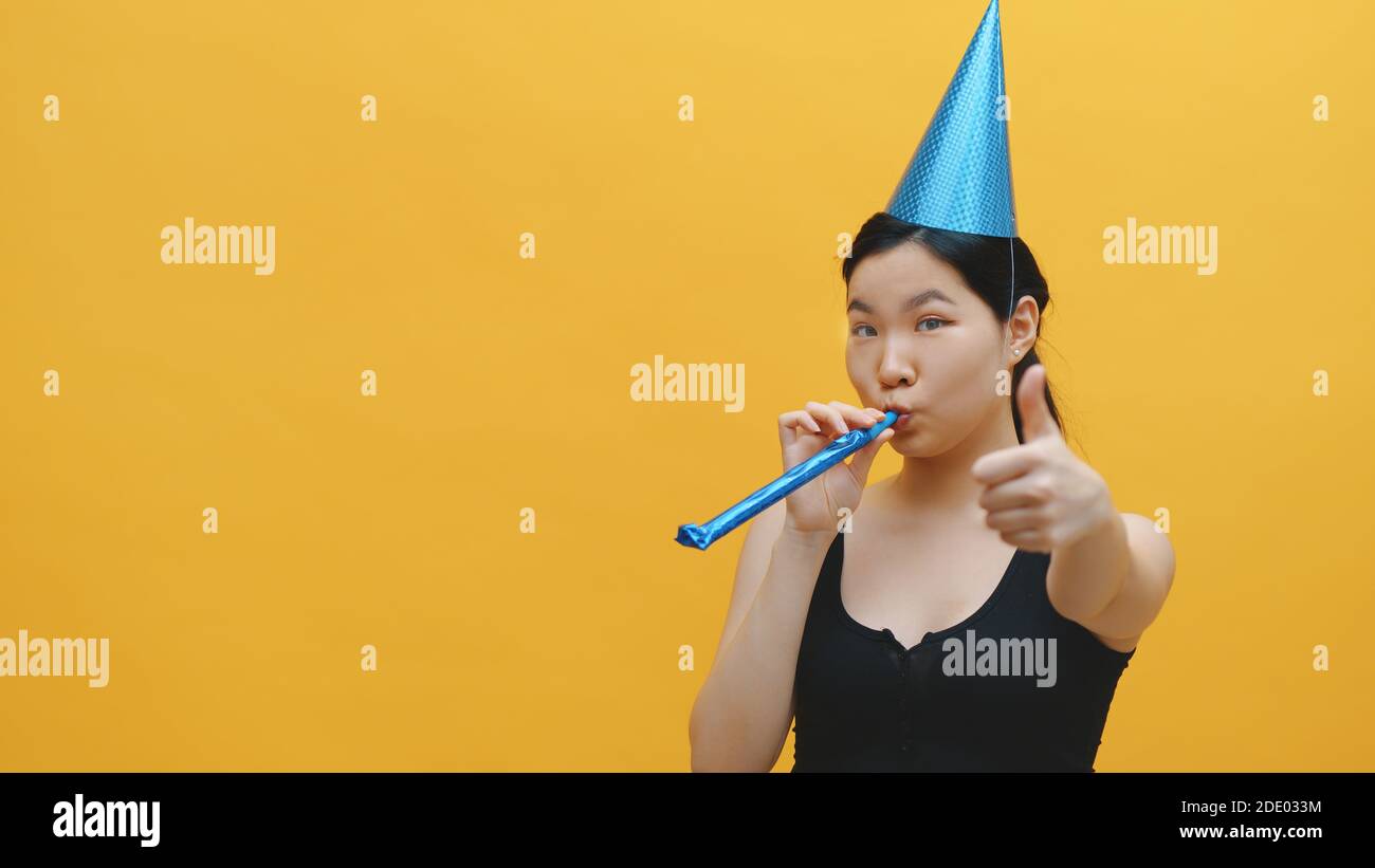 Nette asiatische Frau mit Party Hut weht in die Partei Pfeife und zeigt Daumen nach oben. Party und Feier Konzept. Hochwertige Fotos Stockfoto