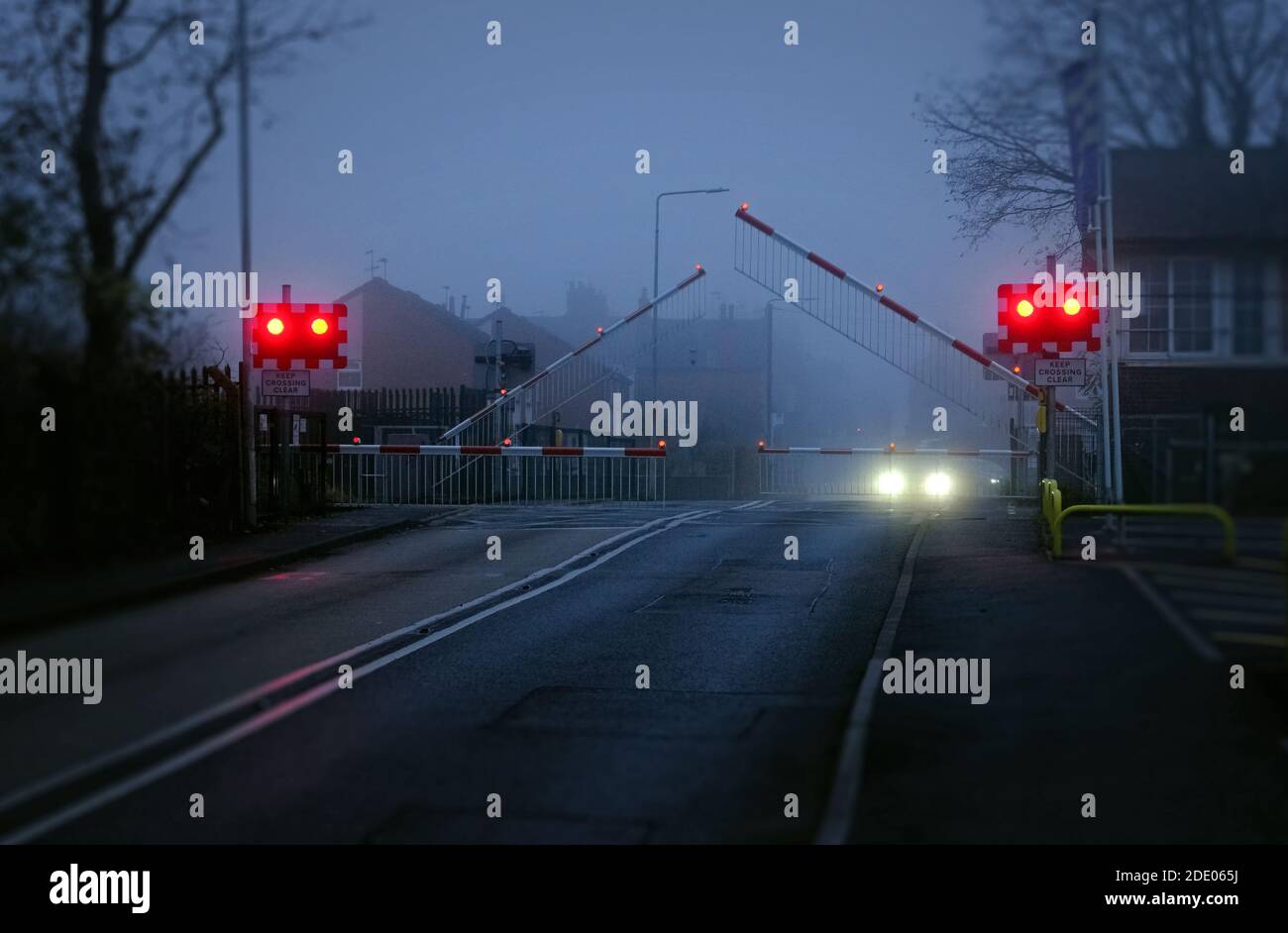 Eine neblige atmosphärische Straße in Bingham, Nottinghamshire, wenn die Barrieren an einem Bahnübergang herunterfallen. Stockfoto