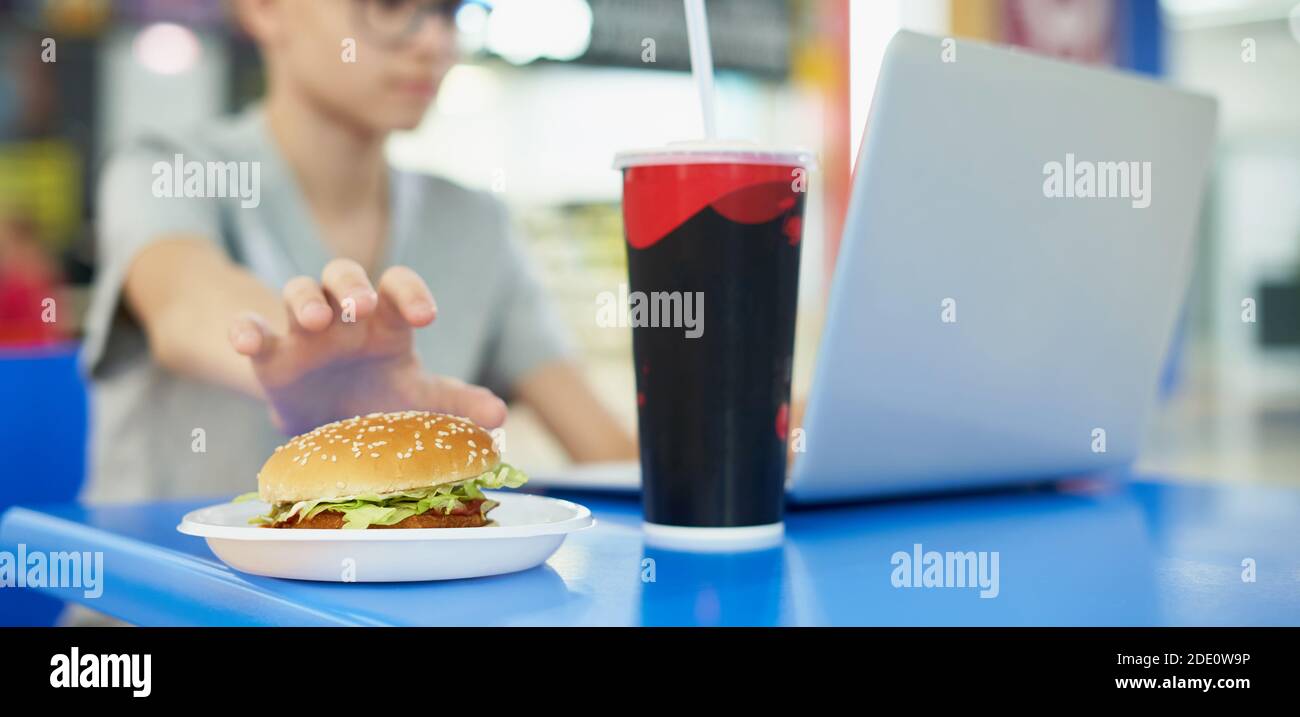 Der Teenager nimmt Burger, während er im Food Court sitzt Eines Einkaufsbummel und nutzt einen Laptop Stockfoto