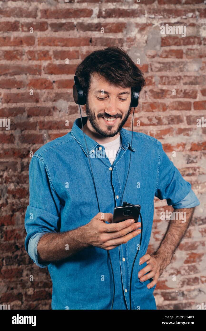 Porträt eines lächelnden jungen Mannes mit Kopfhörer und Handy. Freizeitkleidung und Backsteinwand im Hintergrund. Stockfoto