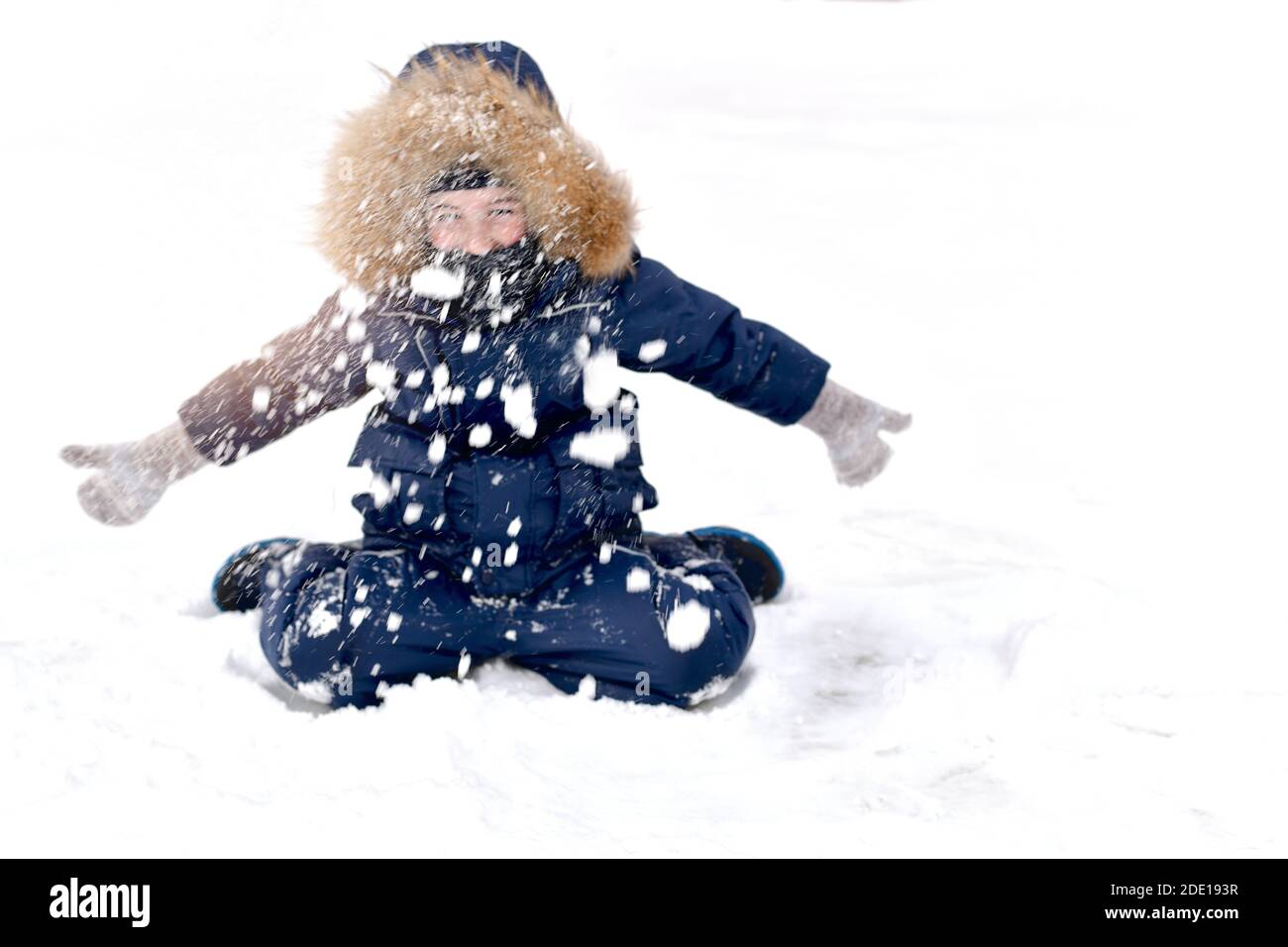 Das Kind ist warm gekleidet, mit einer Pelzhaube. An einem frostigen Wintertag sitzend, mit den Händen in Fäustlingen, wirft er viele gesammelten Schnee hoch, sc Stockfoto