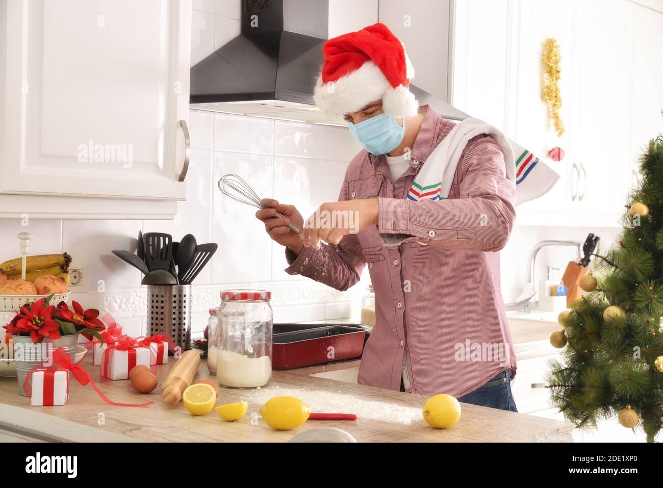 Mann durch covid-19 Kochen ein Dessert mit Zutaten auf beschränkt Die Küchenbank in einem Weihnachtshut und Weihnachten gekleidet Baumhintergrund Stockfoto