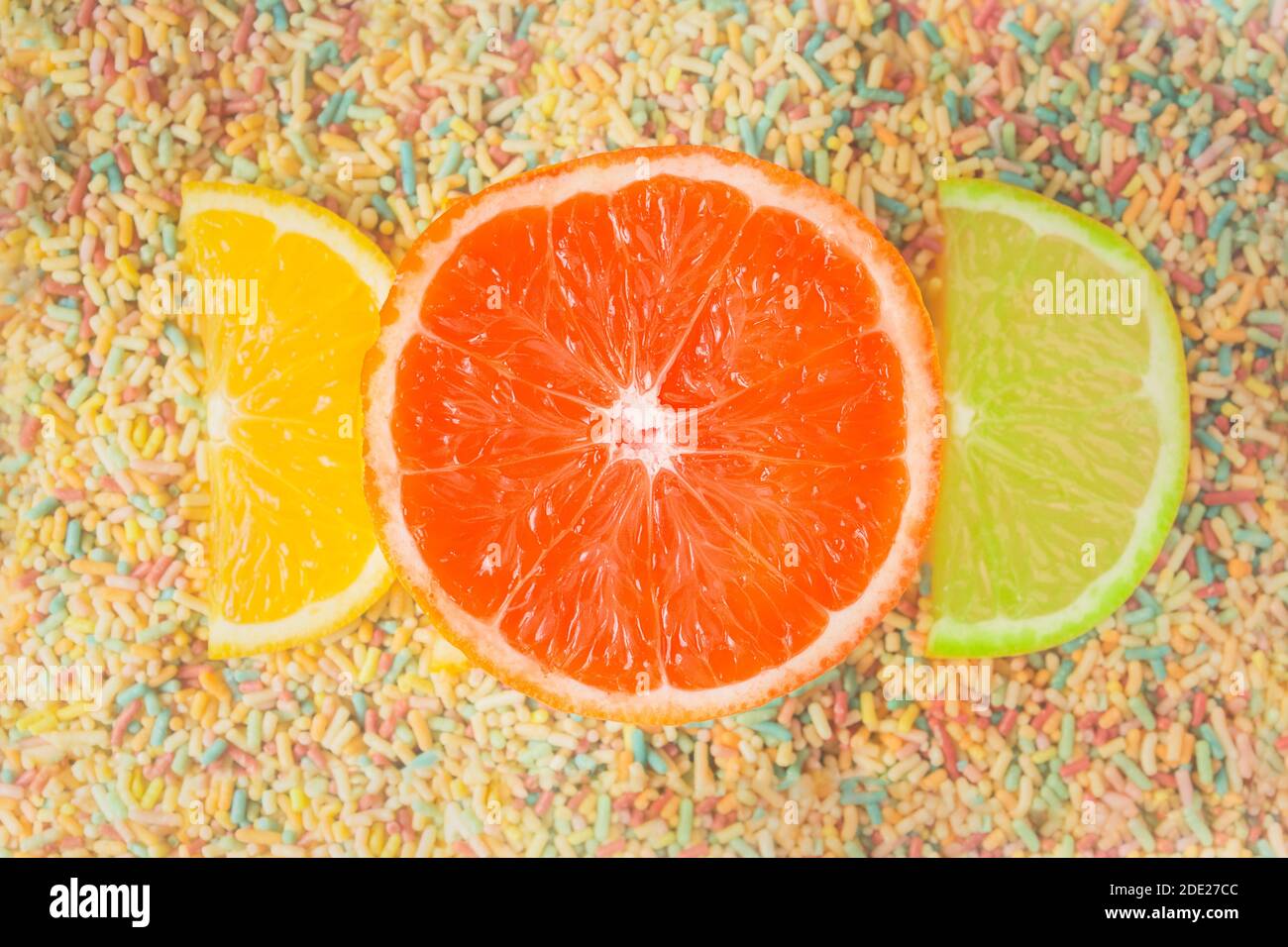 Lustige und bunte Komposition aus halben Scheiben Orange und Limette und einer großen Grapefruit auf einem Hintergrund von farbigen Süßigkeiten. Obst und Zucker Launen. Stockfoto