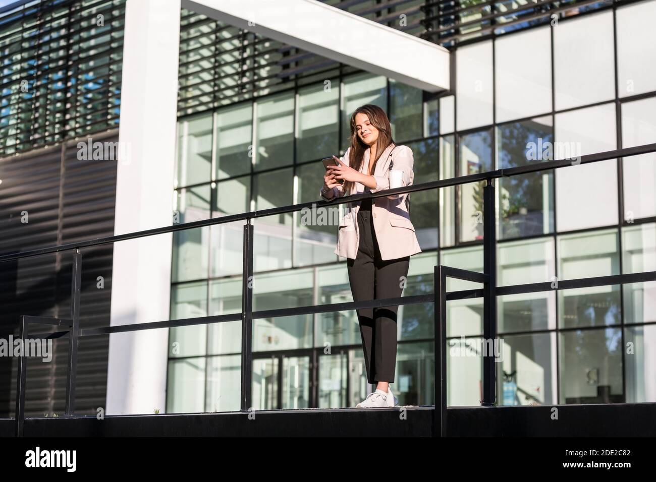 Eine junge, schöne Frau, die sich an einen Zaun lehnt und lächelt, während sie auf ihrem Handy tippt. Sie ist Business-ausgebildet, mit moderner Architektur in der b Stockfoto
