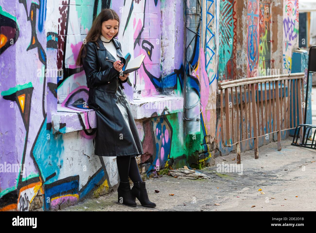 Riga, Lettland - 17. Oktober 2020: Eine junge Frau lehnt sich an eine mit Graffiti besprühte Wand und macht Notizen in einem schwarzen Notizbuch Stockfoto