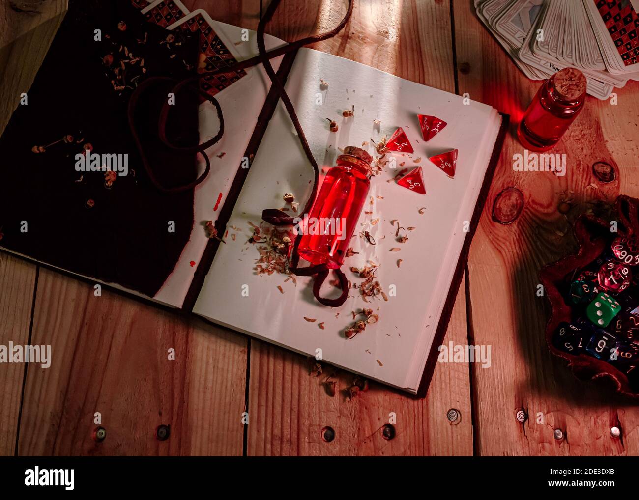 Bild einer Flasche mit roter Flüssigkeit und 4 d4-Würfel auf einem ledergebundenen Tomé. Spielkarten und ein Würfelbeutel voll mit verschiedenen Würfeln auf einem Woo Stockfoto