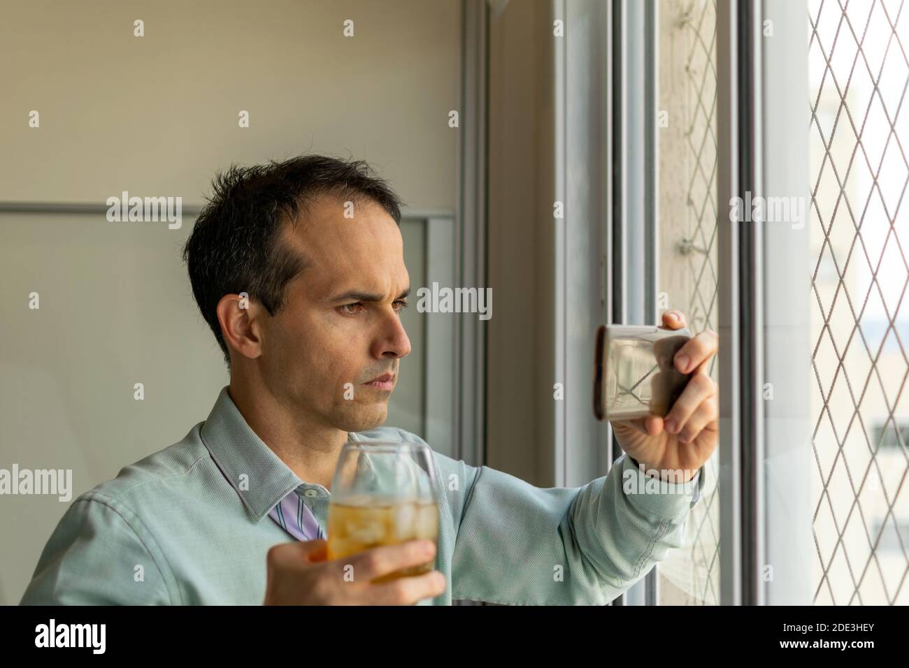 44-jähriger Mann, vor einem Fenster, nimmt einen Whisky und schaut sich Videos auf seinem Smartphone an. Stockfoto