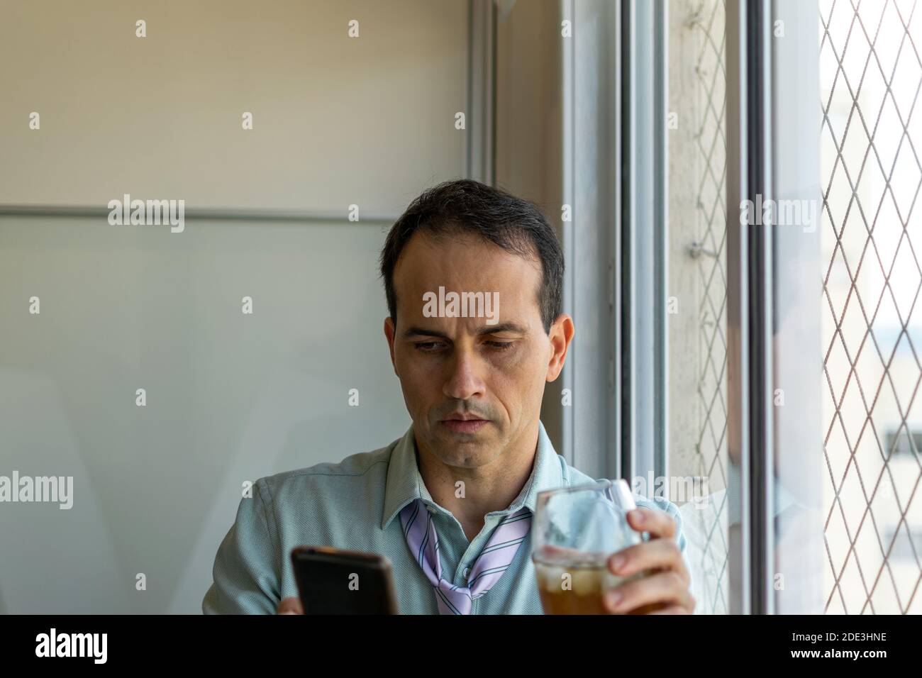 Reifer Mann (44 Jahre alt) hält ein Glas Whiskey und schaut auf sein Smartphone. Stockfoto