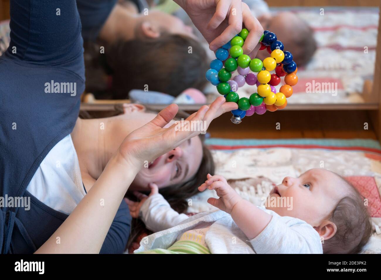 Eine Mutter interagiert und spielt mit ihrer 4 Monate alten Tochter, zeigt ihr ein Spielzeug, wobei beide auf einer Spielmatte mit einem Spiegel dahinter liegen, Großbritannien Stockfoto