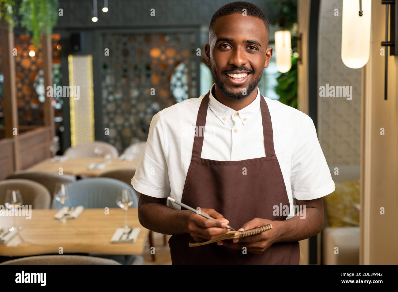 Glücklicher junger Kellner mit toothy Lächeln halten Stift und klein Notepad beim Stehen vor der Kamera im Restaurant und Bestellungen werden entgegengenommen Stockfoto