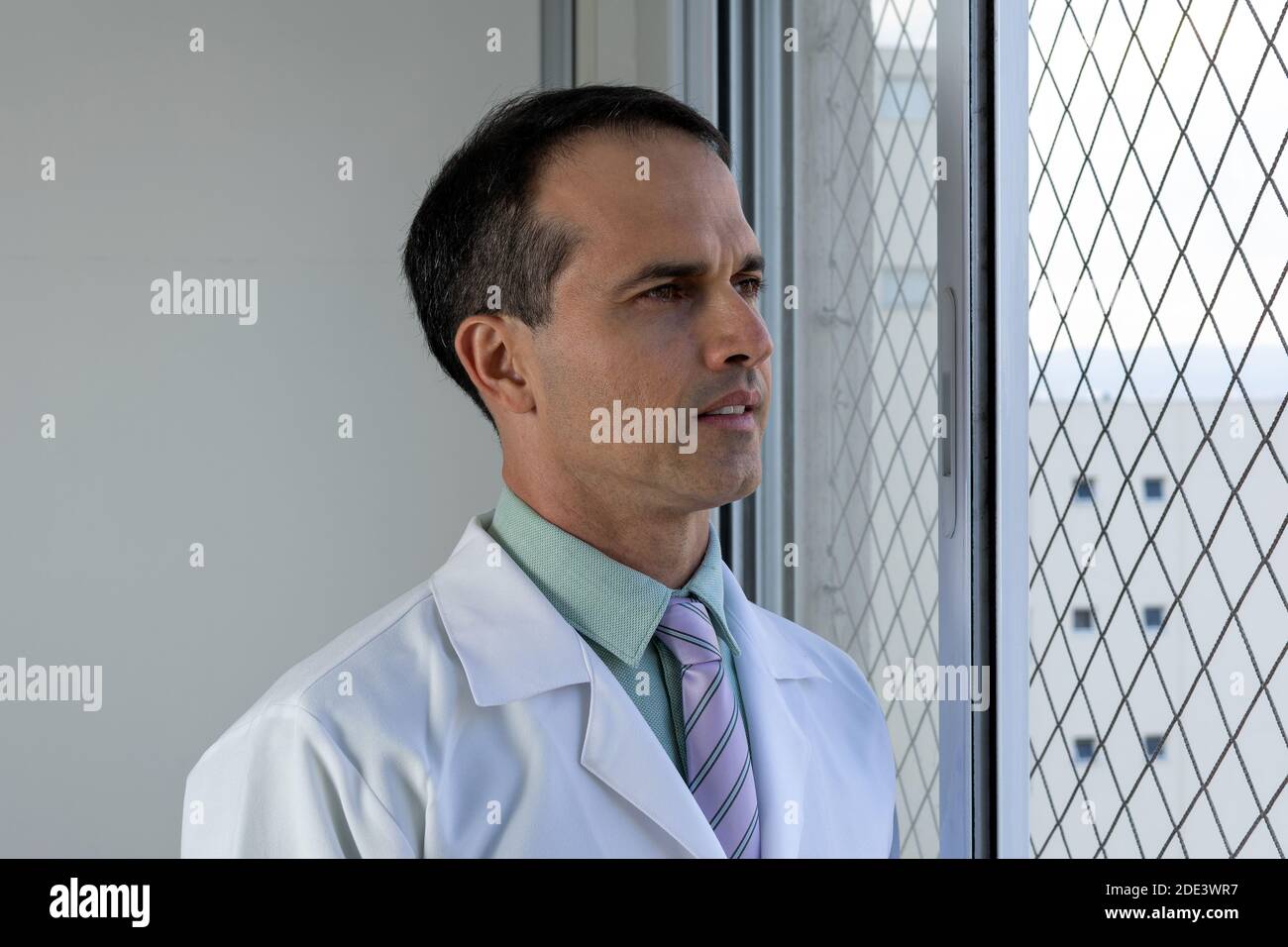 44 Jahre alter Arzt mit weißem Mantel und Krawatte, der auf das Fenster schaut. Stockfoto