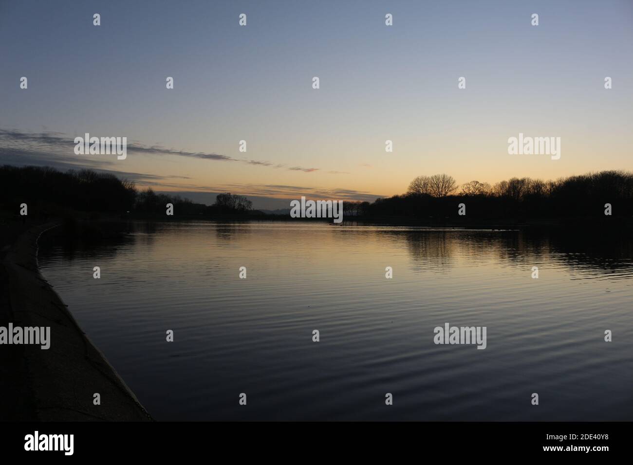 Blick bei Sonnenuntergang mit blauem Himmel mit einigen Wolken am Horizont, die sich an einem kalten Herbsttag auf einem Seewasser spiegeln, intensive orange und dunkle Farben in der Dämmerung Stockfoto