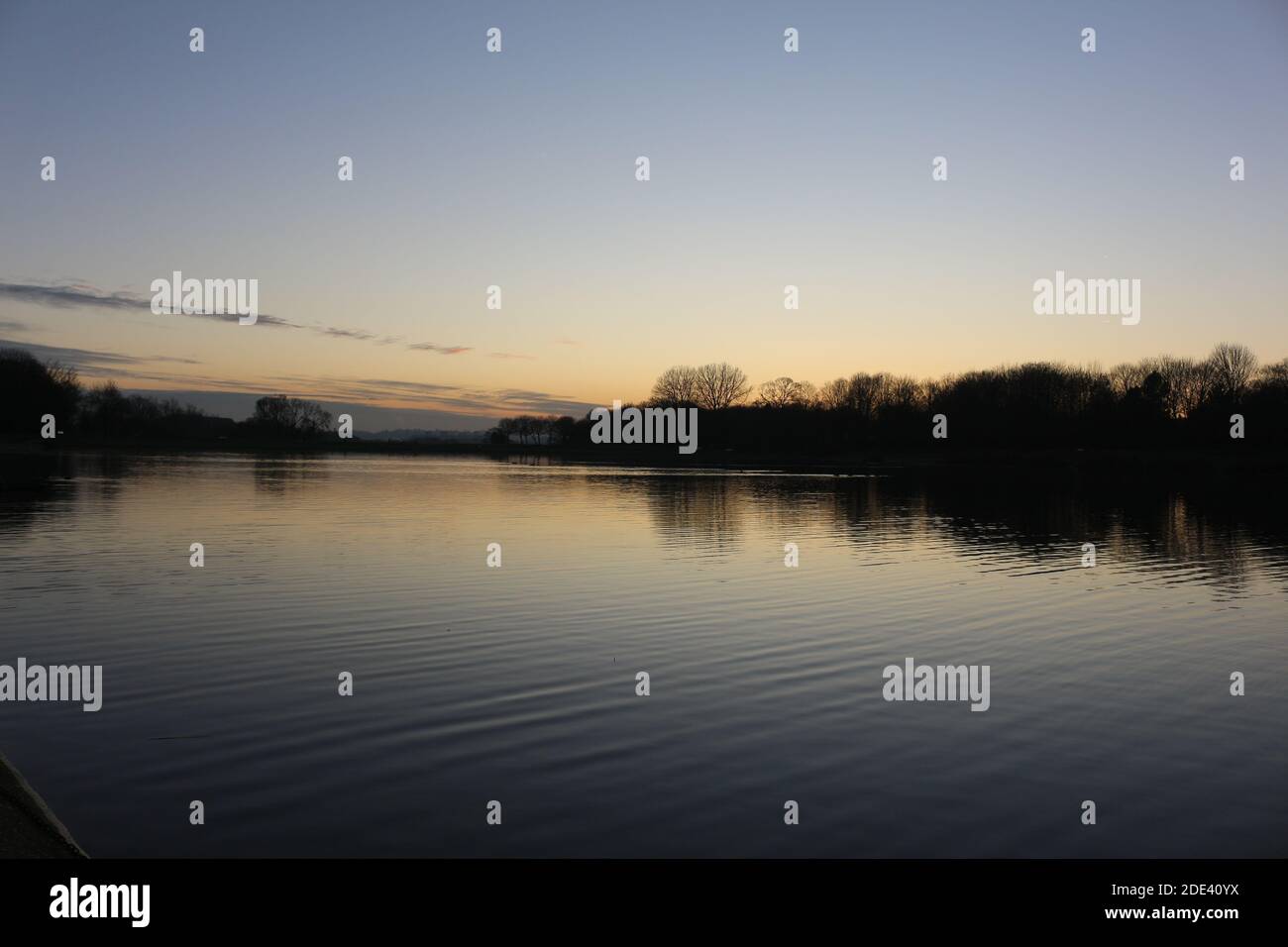 Blick bei Sonnenuntergang mit blauem Himmel mit einigen Wolken am Horizont, die sich an einem kalten Herbsttag auf einem Seewasser spiegeln, intensive orange und dunkle Farben in der Dämmerung Stockfoto