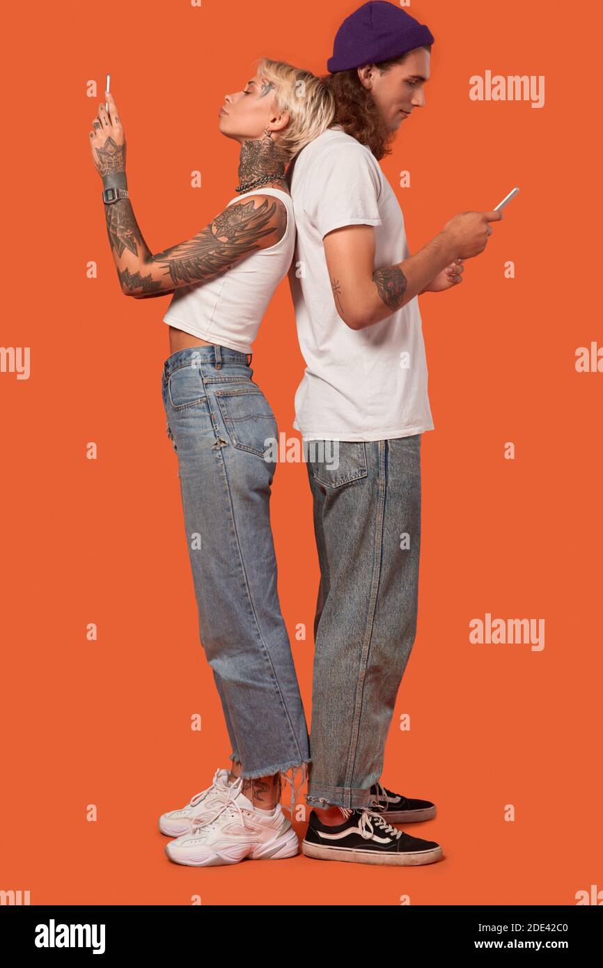 Freistil. Junge tätowierte Paar stehend zurück zu Rücken isoliert auf orange mit Smartphone chatten mit Freund gelangweilt vollen bodu Schuss Stockfoto