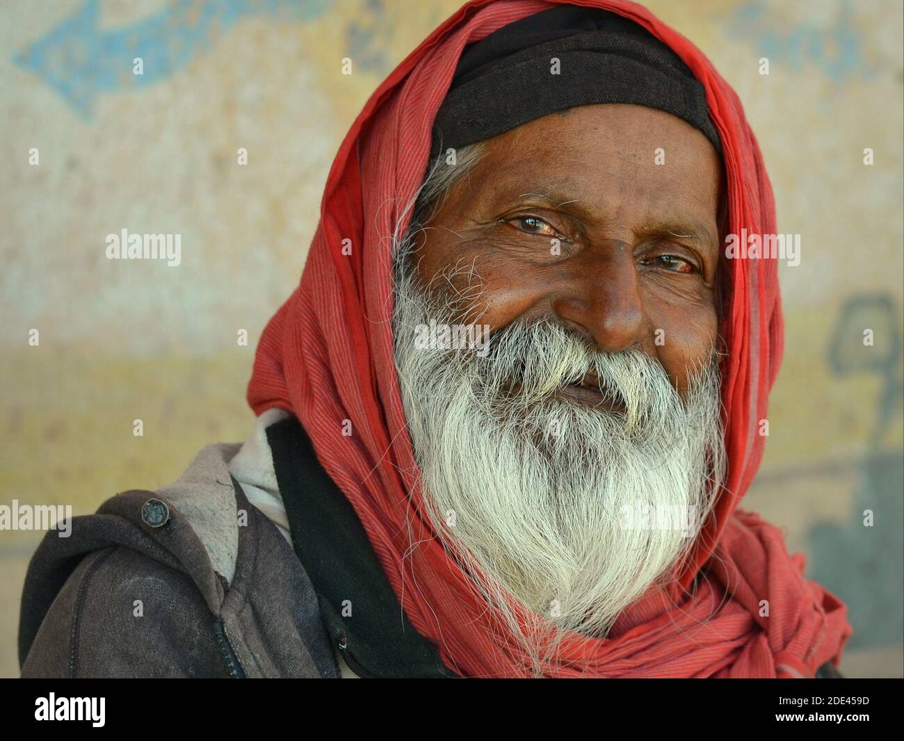 Positiver alter indischer Hindu-Pilger-Großvater mit shampooned weißem Bart und freundlichen Augen bedeckt seinen Kopf mit einem roten Schal und lächelt für die Kamera. Stockfoto