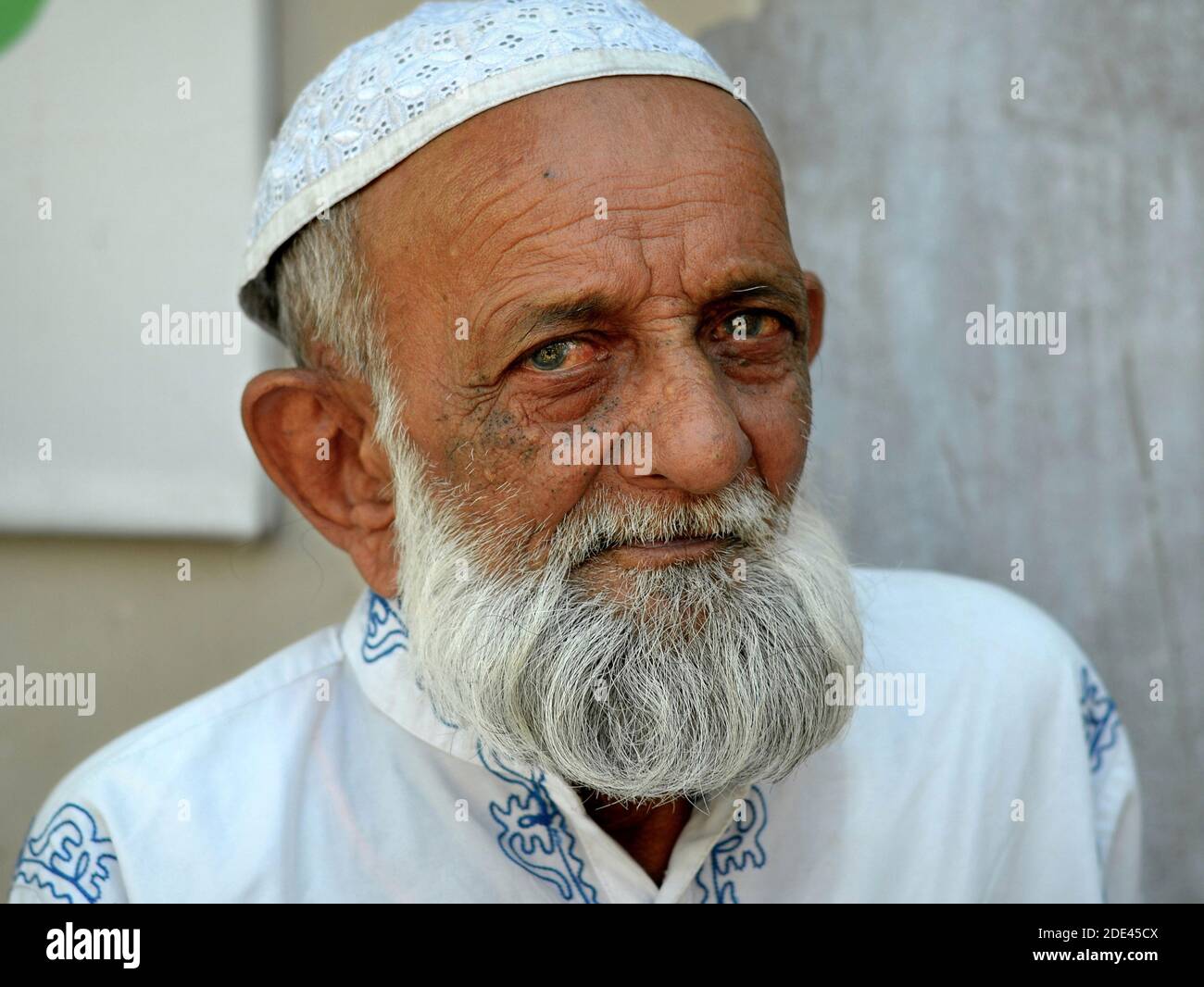Freundlicher älterer indischer Muslim mit islamischem Bart trägt eine weiße islamische Gebetsmütze (Taqiyah) und posiert für die Kamera. Stockfoto