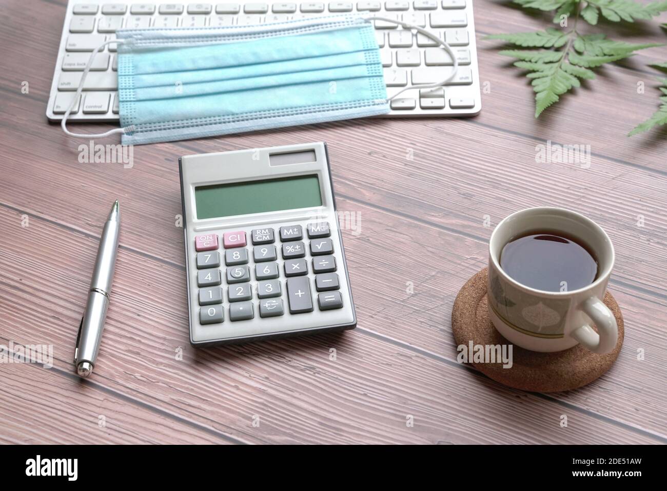 Holztisch mit Tastatur, Gesichtsmaske, schwarzem Kaffee, Stift, Rechner und grünen Pflanzen. Stockfoto