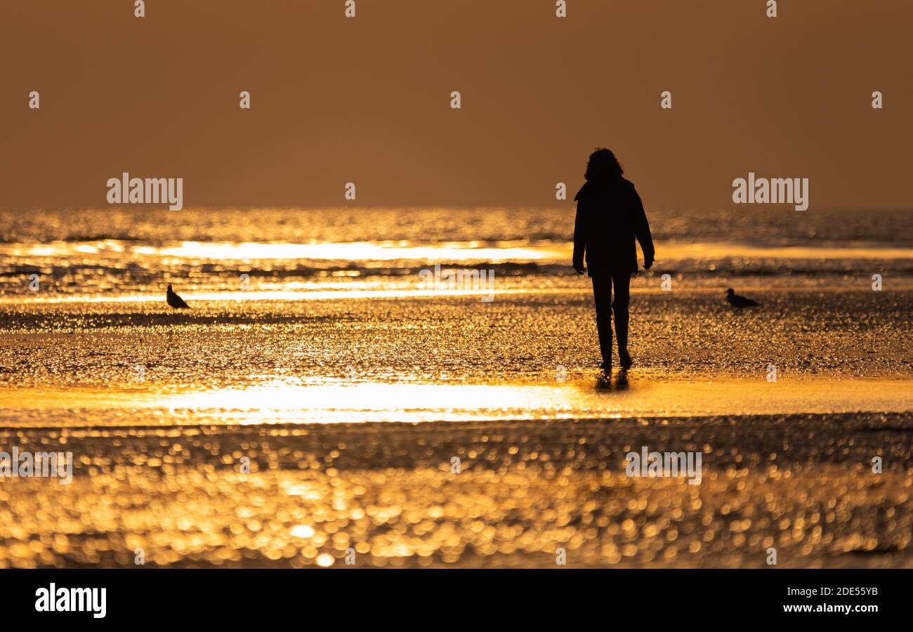 Frau, die am Abend bei Sonnenuntergang alleine an einem Strand spazieren geht, während die Sonne bei Ebbe am Strand reflektiert, in West Sussex, England, Großbritannien. Stockfoto