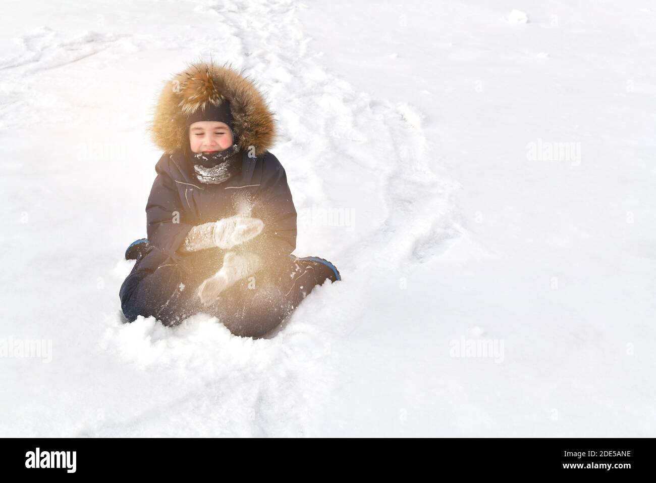 Ein Kind in Kleidern, in einem Daunenjumpsuit, einer Wollmütze und einer Kapuze mit Fell, das im Winter auf dem Schnee sitzt, schüttelt den Schnee von den Fäustlingen ab und klopft sein ha Stockfoto