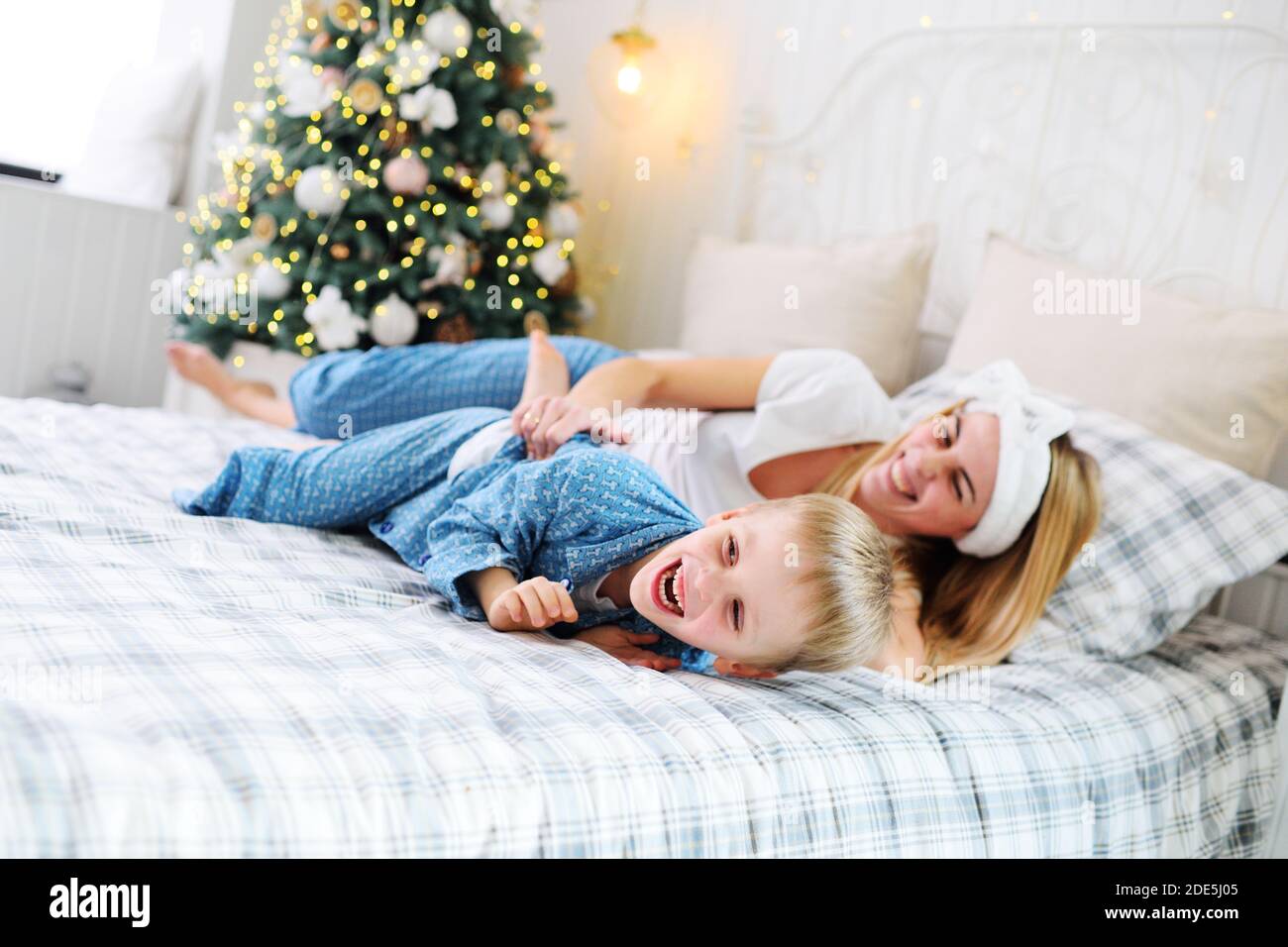 Die junge Mutter lacht und kitzelt ihren kleinen Sohn im Pyjama, während sie auf dem Bett vor dem Hintergrund eines Weihnachtsbaums liegt. Stockfoto