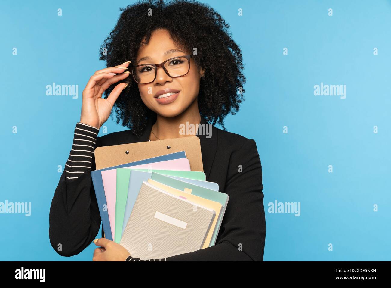 Glückliche Lehrerin oder Geschäftsfrau in Blazer, tragen Sie eine Brille, halten Notebooks, Dokumente und Ordner, lächelnd Blick auf Kamera isoliert auf blauen Studio Wal Stockfoto