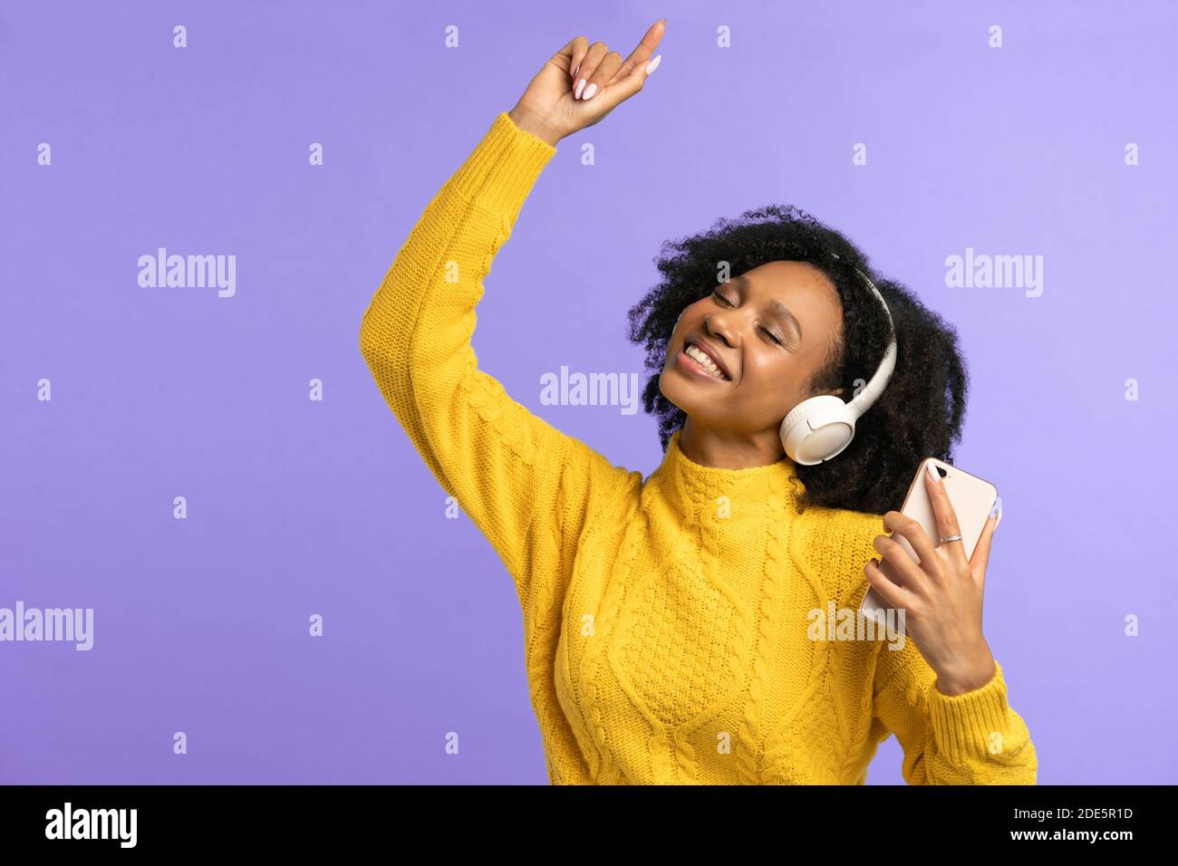 Energiegeladene Mixed Race Frau singt mit, lächelt breit, trägt kabellose Kopfhörer, hat gute Laune, isoliert auf violettem Hintergrund. Fröhliche dunkelhäutige Locke Stockfoto