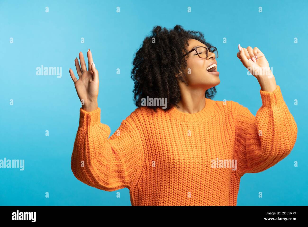 Lustige dunkelhäutige Millennial Frau mit lockigem Haar in guter Stimmung singt ihr Lieblingslied, tanzen, tragen Hipster Oversize orange Pullover, isoliert auf s Stockfoto