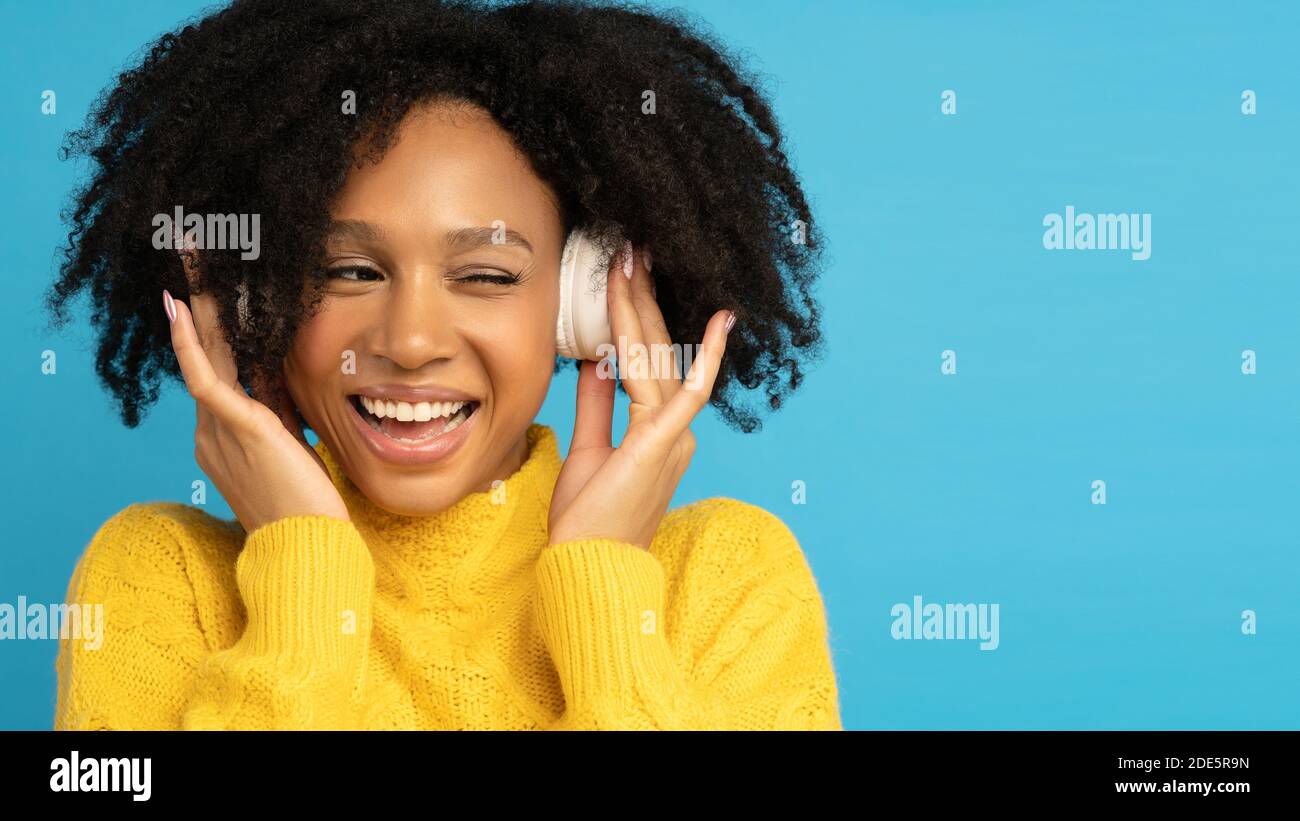 Lächelnde Mischling Frau im gelben Jumper berührt kabellose Kopfhörer, hört Musik, hat gute Laune, zwinkert und schaut auf leeren Raum für Werbung Stockfoto