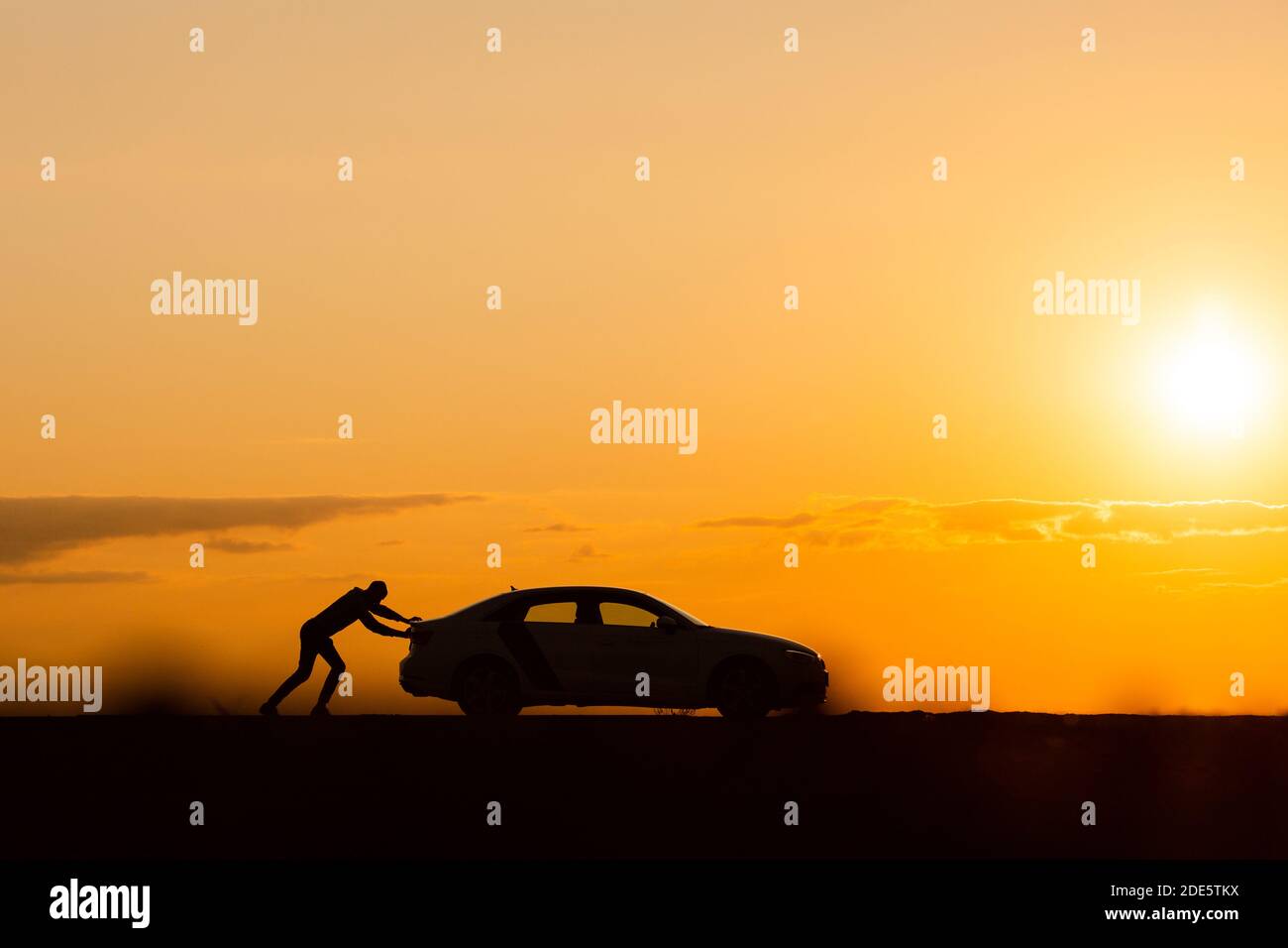 Transportprobleme. Silhouette eines Mannes Fahrer schiebt sein Auto entlang auf einer leeren Straße nach dem Zusammenbruch bei Sonnenuntergang, Kopierraum, Seitenansicht. Stockfoto