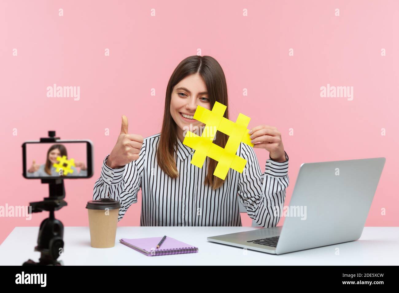 Positiv lächelnde Bloggerin, die Videobotschaft für ihre Follower auf dem Smartphone aufzeichnet und ein gelbes Hashtag-Schild zeigt, das nach einer Bewertung des Beitrags fragt. Studio im Innenbereich Stockfoto