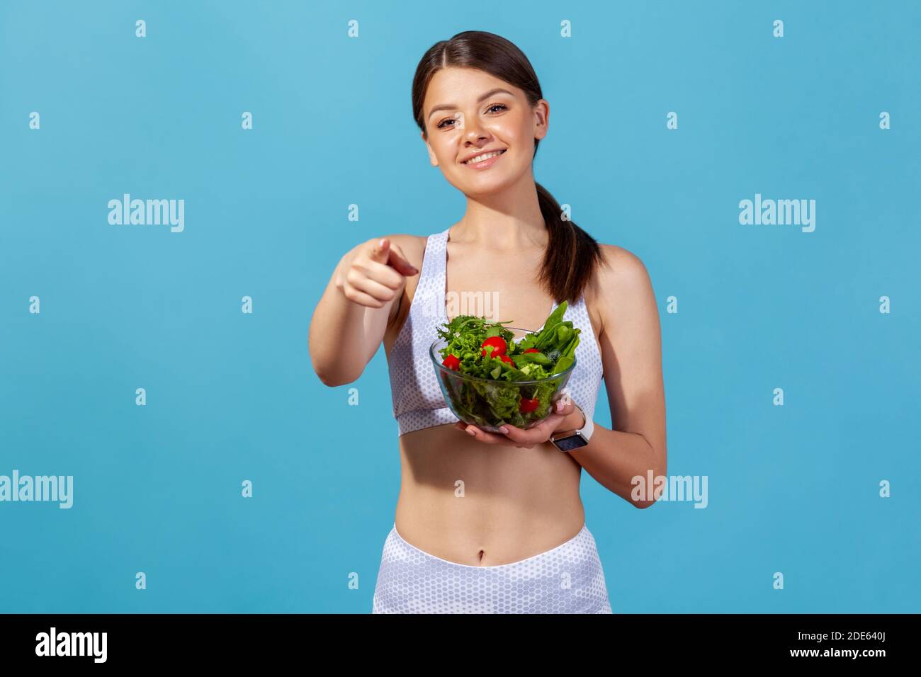 Lächelnde Frau ist weiße Sportswear mit schlanken Körper zeigt Finger auf Sie halten Schüssel mit Gemüsesalat, was darauf hindeutet, Ernährung. Studio im Innenbereich Stockfoto