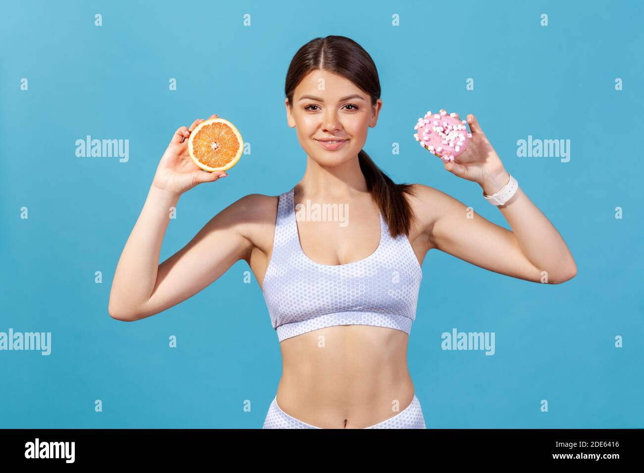 Gesunde schlanke Frau in Sportswear hält Orange und Donut in den Händen, die Wahl zwischen gesunden und Junk-Food-Ernährung. Innenaufnahme des Studios isoliert Stockfoto