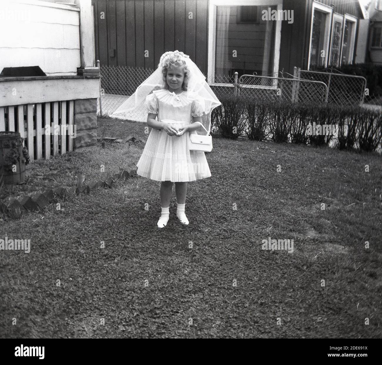 1960s, Historical, USA, ein süßes kleines Mädchen, das als Brautjungfer verkleidet ist und in einem Vorgarten für ihr Foto steht. Weibliche Brautjungfern werden manchmal als Blumenmädchen bezeichnet. Stockfoto